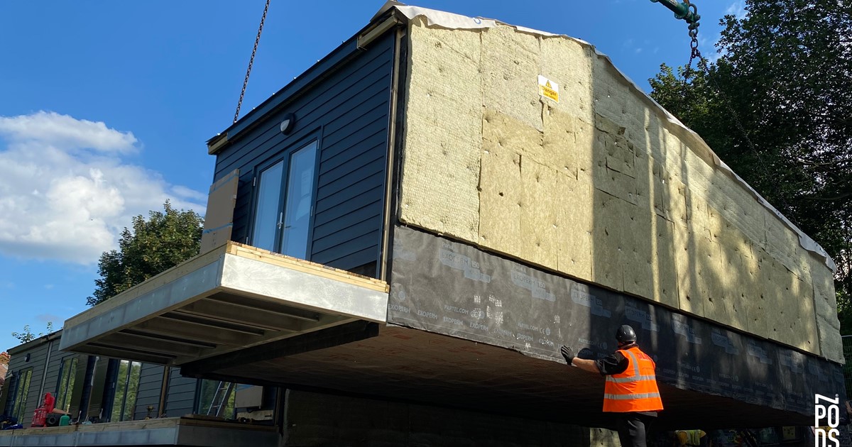 A modular house is lifted into place on a crane. There is a worker in an orange high vis vest helping with the task, which is taking place on a bright, sunny, blue sky day.