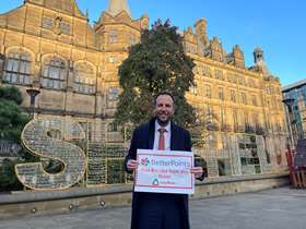 Councillor Ben Miskell stands in front of Sheffield Town Hall holding a poster advertising the Christmas Public Transport Ticket Offer, behind him is the Sheffield sign made up of Christmas lights.