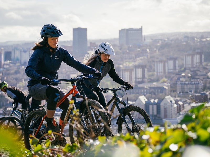 Two mountain bikers ride through one of the trail routes at Parkwood Springs with Sheffield's Arts Tower and the Royal Hallamshire Hospital part of the cityscape in the background.