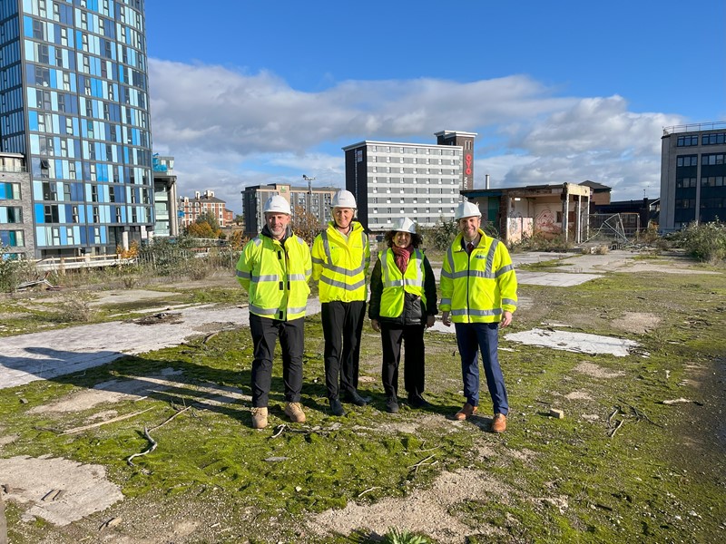 Four people, all wearing high visibility jackets and hard hats stand on waste land within the Castlegate development behind them is a high rise residential block and there is also a blue sky with some clouds in the distance.
