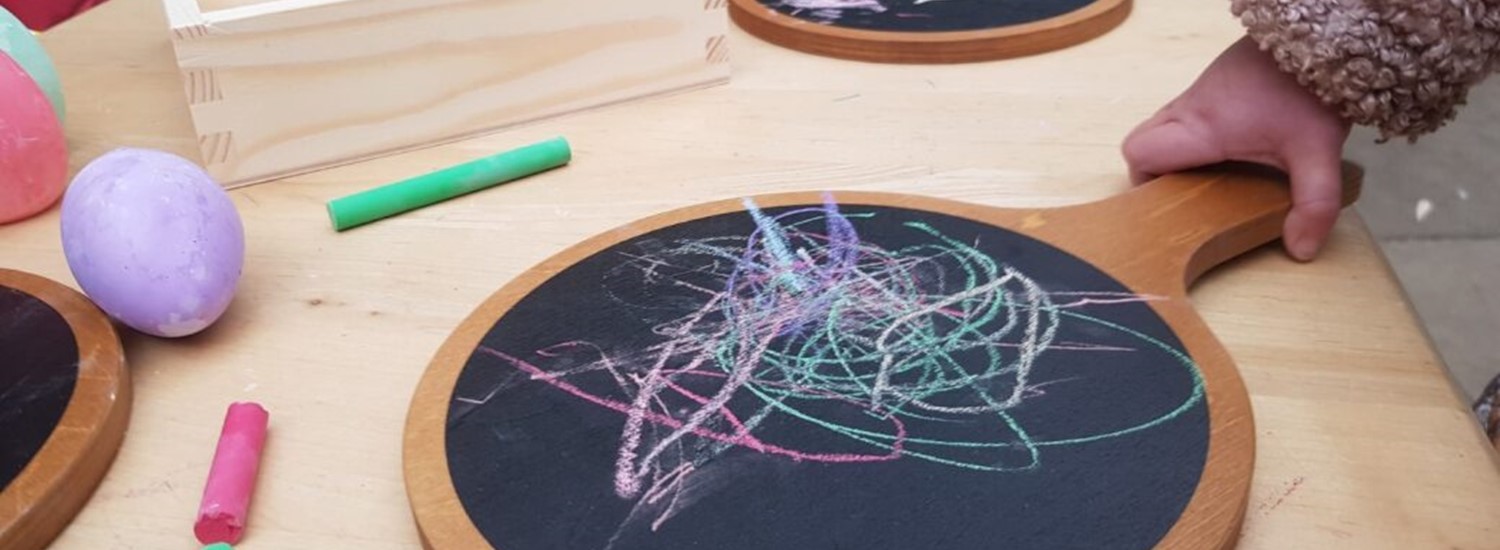 A child holds a chalk board in their hand on which they have doodled while working at a table, with other chalk boards, children and chalk can be seen in the background