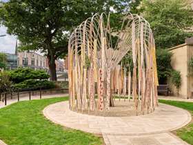Stainless steel willow tree sculpture in paved area with gardens surrounding. Ribbons are attached to the branches of the tree