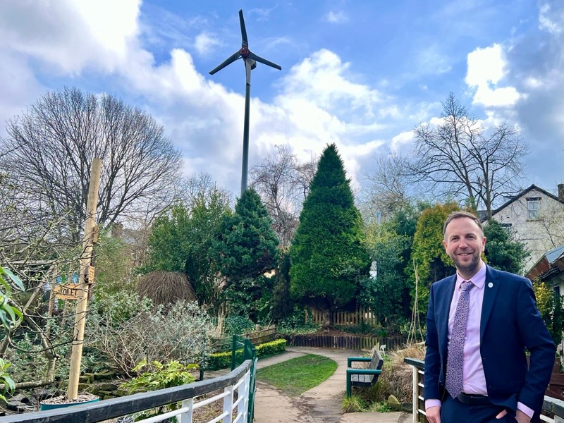 Cllr Ben Miskell stands in the foreground with a wind turbine in the background, scattered throughout the picture are a number of trees and a path disappears off into the distance