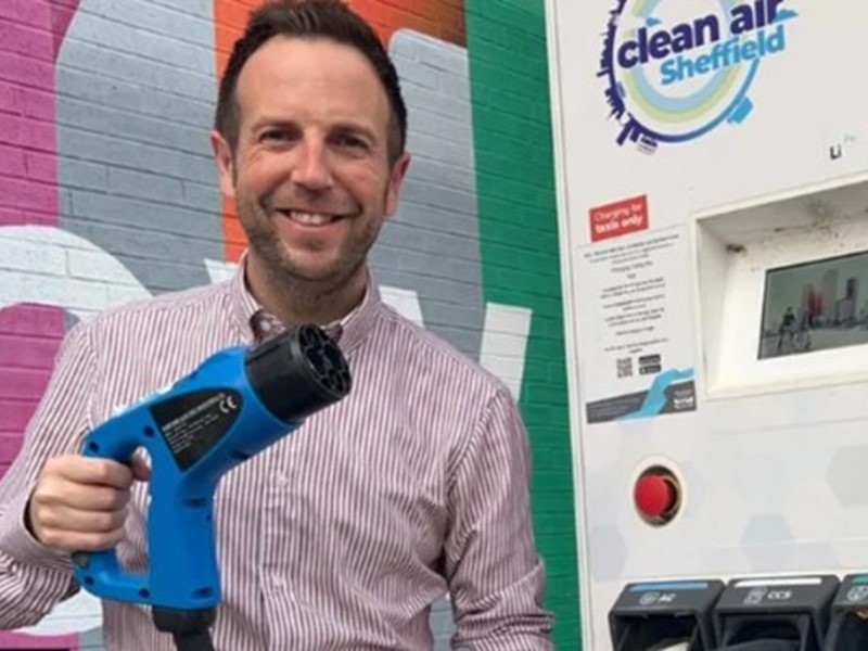 Cllr Ben Miskell standing next to a white electric vehicle charging point, he is holding the plug that goes into the cars in his right hand