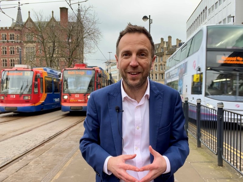 Cllr Ben Miskell stands in Sheffield city centre with two trams parked up behind him on his right hand side and a double decker bus on his left hand side