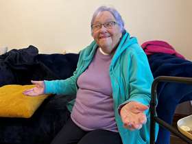 Pat, smiling with her arms raised slightly at her sides sitting on her sofa