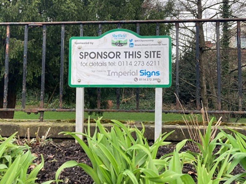 A blank, unused sponsorship sign stands among some grass in front of a metal railing with trees behind