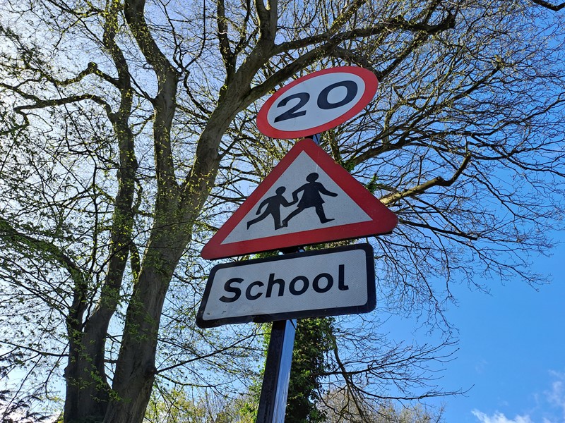 A 20mph sign, children crossing sign and school sign adorn a signpost, with a tree in the background on a sunny day