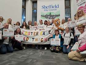 Sheffield City Council fostering service staff, one dressed as a teddy bear, and foster carers pose with a Foster Care Fortnight banner in front of a Sheffield City Council lgo