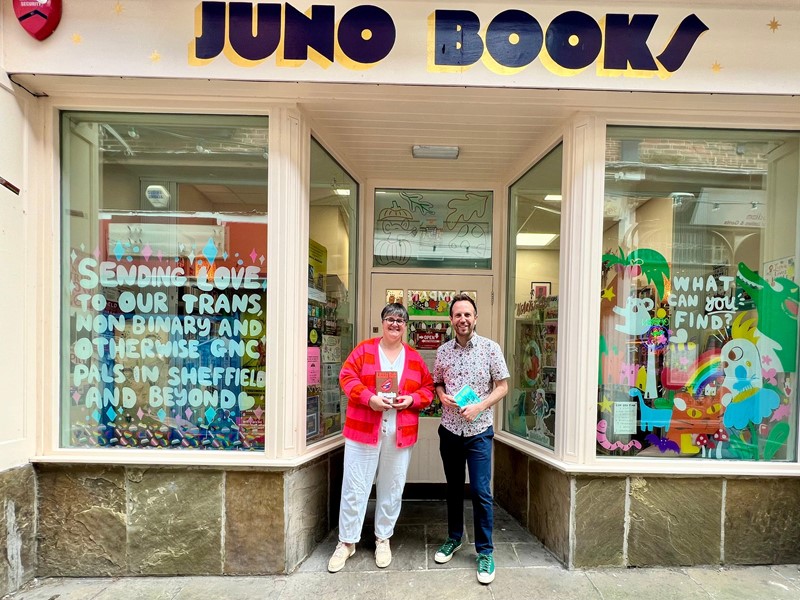 Cllr Ben Miskell and the owner of Juno Books stand in the entrance of a shop, the sign above them says 'Juno Books' and they have windows on either side of them, both windows have got white writing on