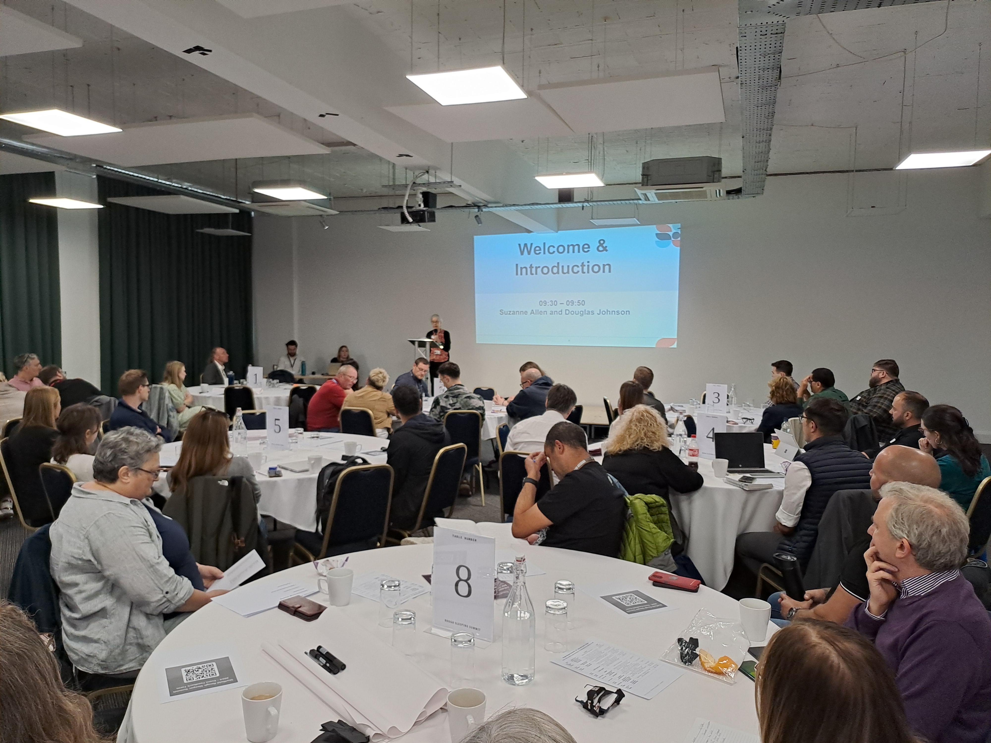 A room full of people sat at tables listen to a speaker in front of a digital screen welcoming them to an event