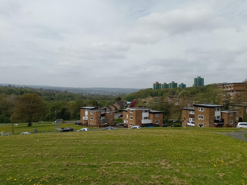 An area of grass with housing behind and high rise blocks in the background on a cloudy day
