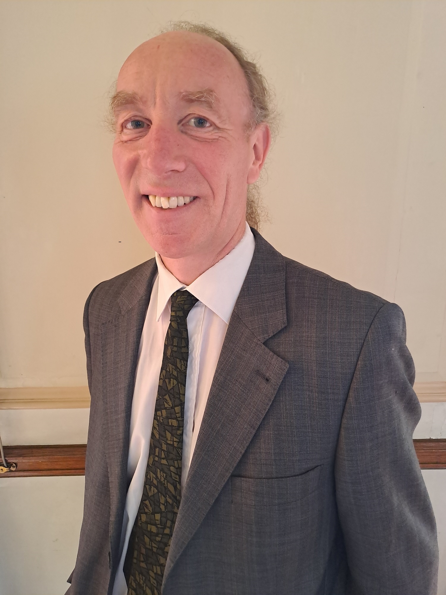 Cllr Douglas Johnson smiles in front of a bright coloured wall, wearing a grey suit, white shirt and green tie