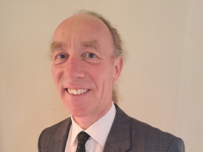 Cllr Douglas Johnson smiles in front of a bright coloured wall, wearing a grey suit, white shirt and green tie