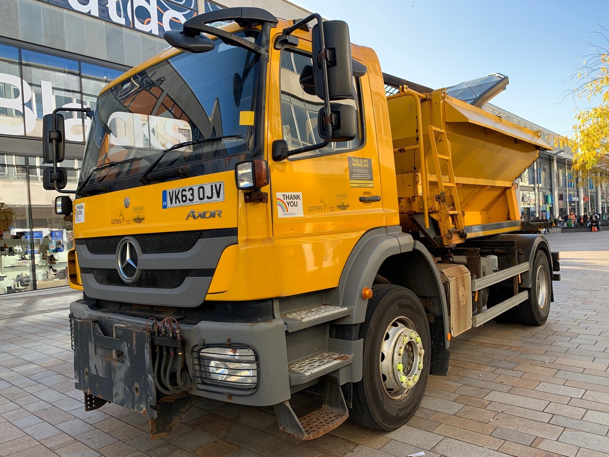 Image shows a side-on picture of a large yellow gritter lorry on a road against a backdrop of a blue sky