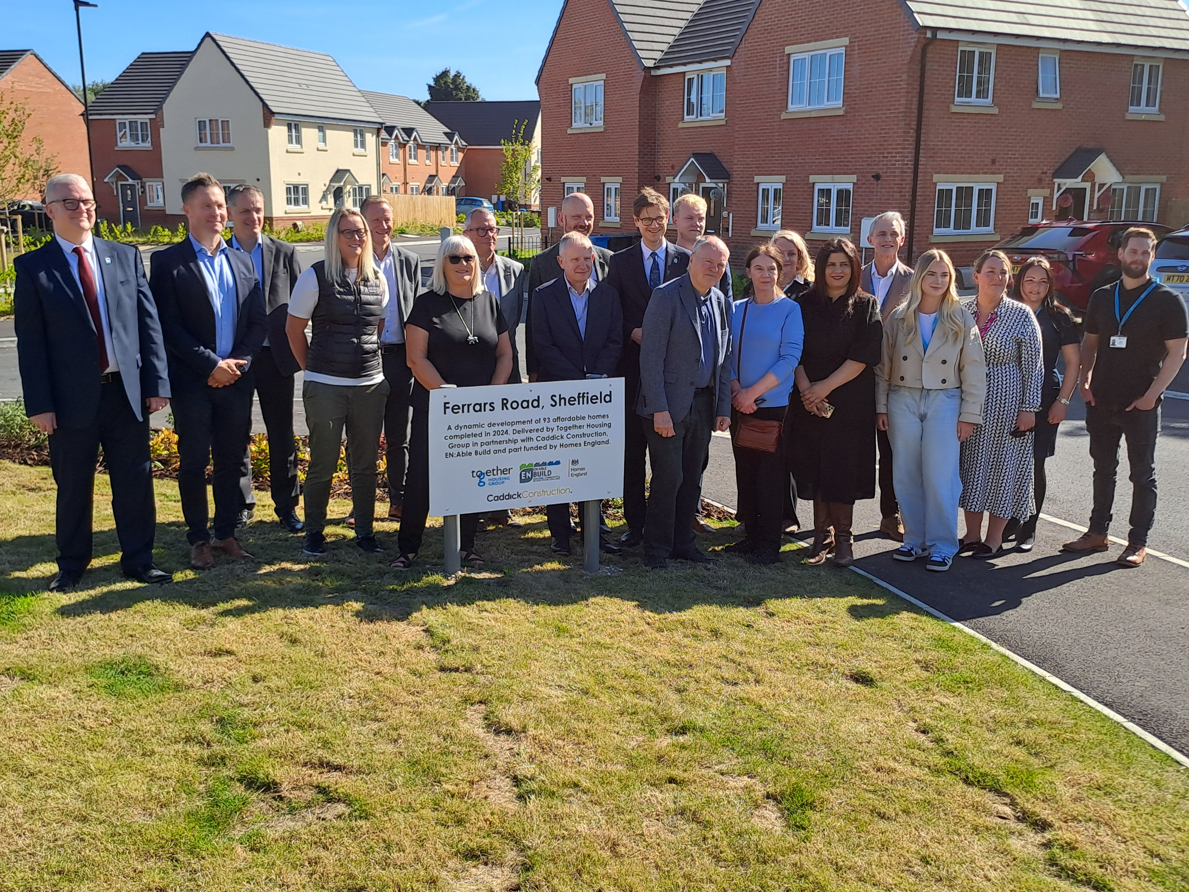 Councillors, council officers, partners and politicians smile while stood across grass and a pavement behind a sign for a new housing development on a sunny day