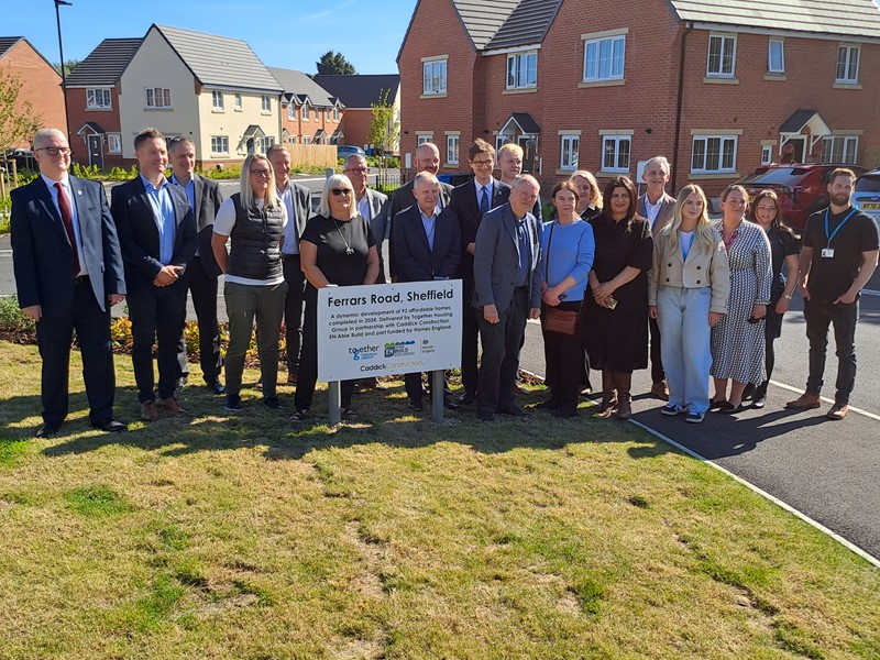 Councillors, council officers, partners and politicians smile while stood across grass and a pavement behind a sign for a new housing development on a sunny day