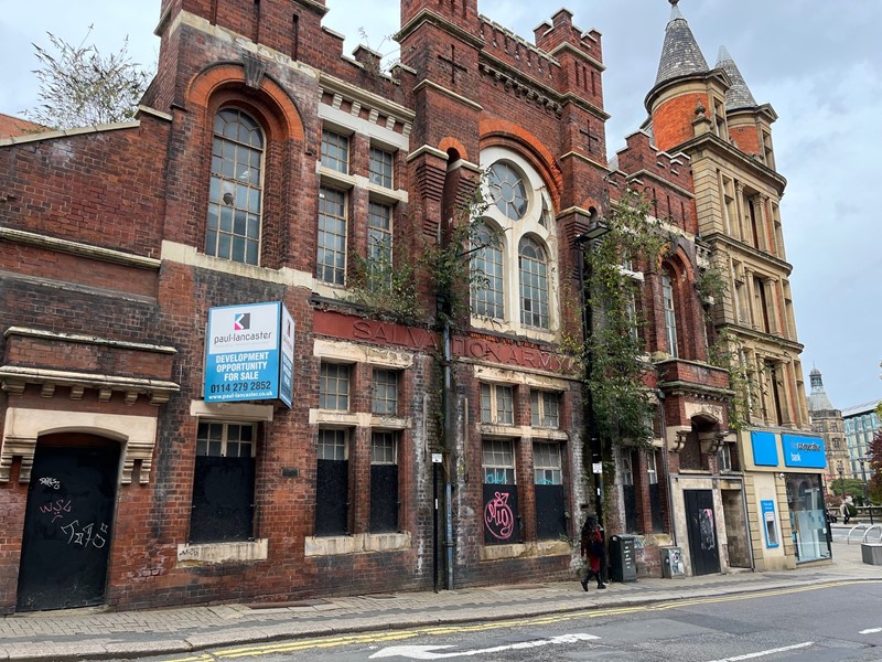 The red-brick frontage of the former Salvation Army Citadel building in Sheffield city centre, the building shows plants growing through the front structure as a dark hair woman walks in front of it.