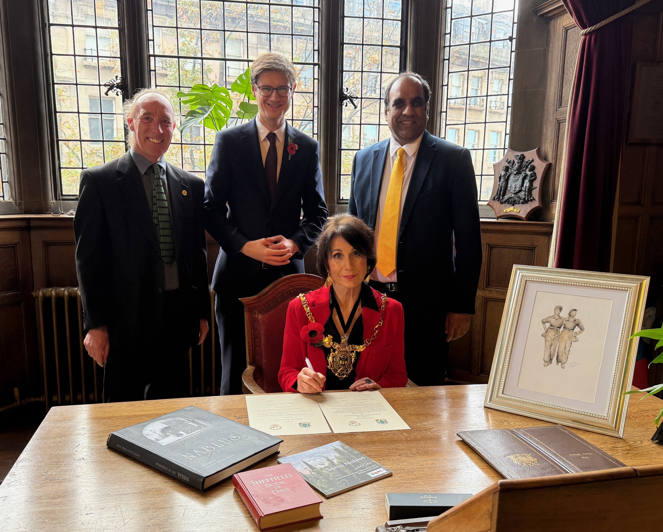 Cllr Johnson, Cllr Hunt and Cllr Mohammad standing behind the Lord Mayor who is sitting at a desk with the document to be signed and books about Sheffield and Nablus laid out