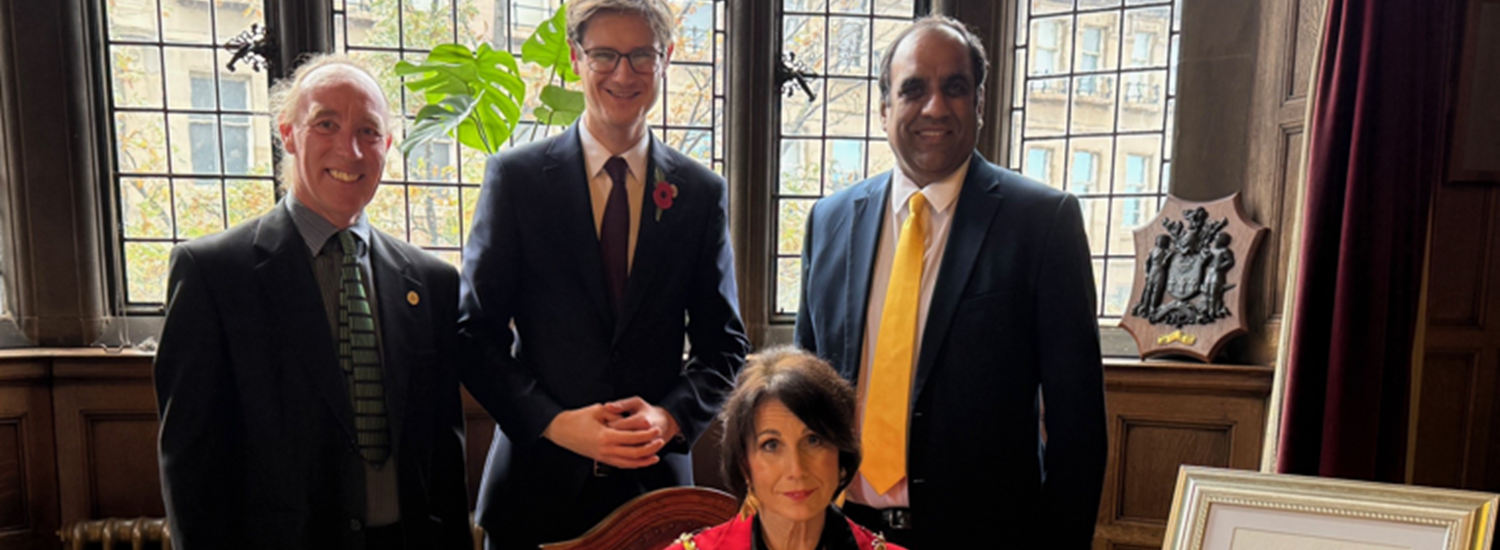 Cllr Johnson, Cllr Hunt and Cllr Mohammad standing behind the Lord Mayor who is sitting at a desk with the document to be signed and books about Sheffield and Nablus laid out