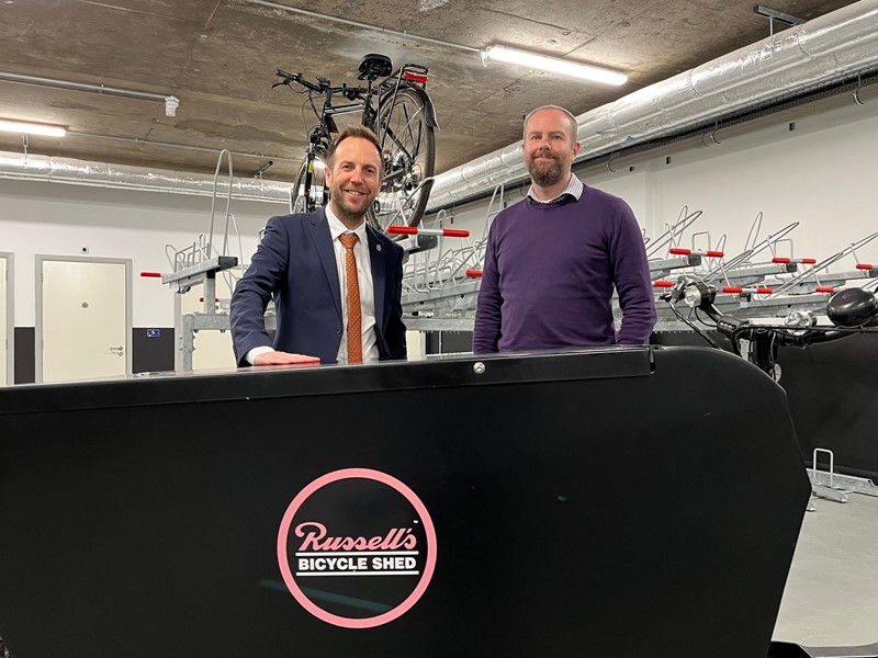 Cllr Ben Miskell, on the left, and Russell Cutts, on the right, stand in the city cycle hub behind a black box with 'Russell's Cycle's' written on it