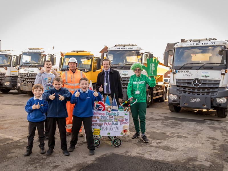 A picture of children and adults stood in front of a line of white and orange gritters
