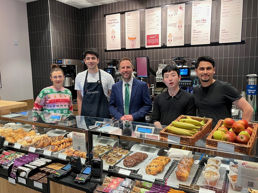 Cllr Ben Miskell stands in the middle of a line of staff members at the new Pret A Manger in Sheffield city centre, the group are stood behind the counter with food products in front of them.