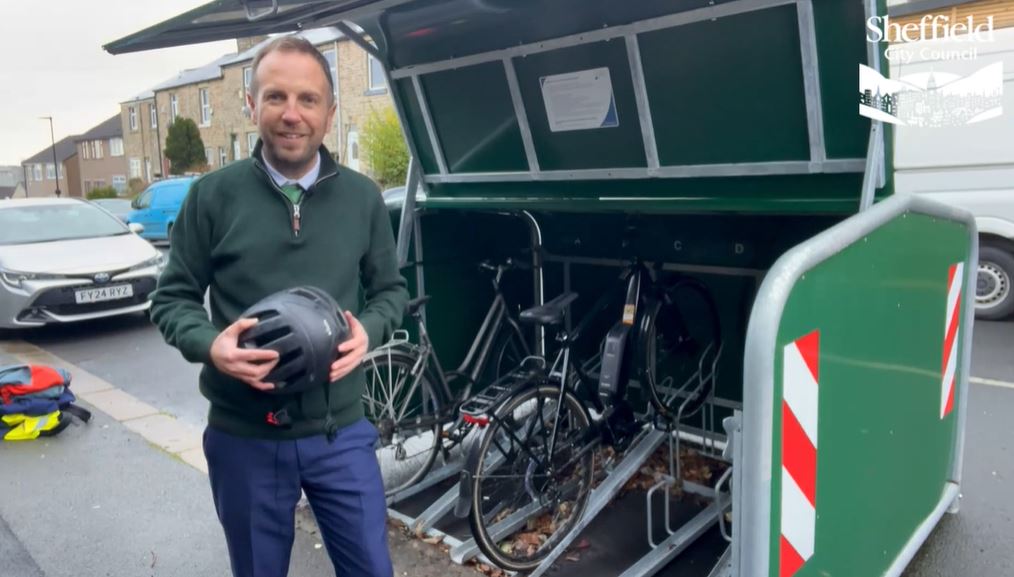 Councillor Ben Miskell stands in front of of an open cycle hangar on a busy street, with cars in the background. There are bikes in the hangar, and information is fixed to the inside of the hangar lid.
