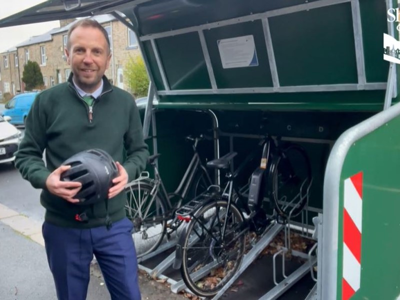 Councillor Ben Miskell stands in front of of an open cycle hangar on a busy street, with cars in the background. There are bikes in the hangar, and information is fixed to the inside of the hangar lid.